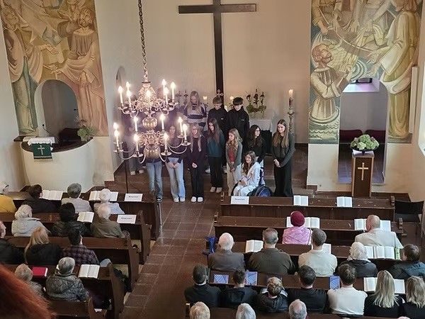 Eine Gruppe von Menschen steht in einer Kirche, mit einem großen Kreuz und einer Kronleuchter im Hintergrund. Die Gemeinde sitzt auf Kirchenbänken, und einige lesen aus offenen Büchern.