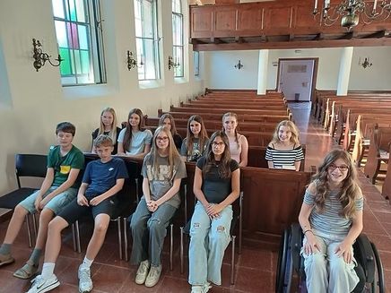 Eine Gruppe junger Menschen, sowohl Jungen als auch Mädchen, sitzen auf einer Kirchenbank und lächeln für ein Foto. Die Kirche hat Holzbänke, Buntglasfenster und eine Kronleuchter.