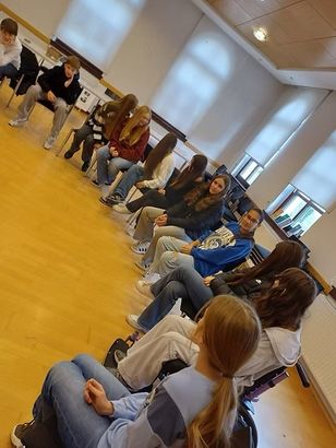 Eine Gruppe junger Menschen sitzt in einem Raum auf Stühlen. Sie tragen Freizeitkleidung und scheinen in einer Diskussion engagiert zu sein. Der Raum hat Holzböden und weiße Wände.
