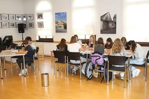 Eine Gruppe von Schülern sitzt um Tische in einem Klassenzimmer herum und führt eine Diskussion. Der Raum ist gut mit natürlichem Licht beleuchtet, und es hängen gerahmte Bilder an der Wand. Ein Mülleimer steht in der Nähe der Tische.