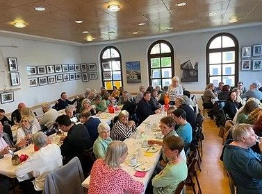 Eine große Gruppe von Menschen sitzt an langen Tischen in einem Speisesaal, einige unterhalten sich, mit gerahmten Bildern an der Wand und heller Beleuchtung.