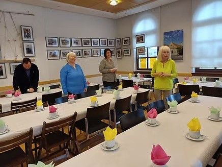 Vier Frauen in einem Raum mit langen Tischen, die für eine Mahlzeit gedeckt sind. Eine Frau spricht, während die anderen zuhören. Der Raum hat gerahmte Bilder an den Wänden und eine helle Atmosphäre.