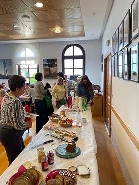 Eine Gruppe von Menschen versammelt sich um einen langen Tisch mit verschiedenen Speisen darauf. Der Tisch ist mit einem weißen Tuch bedeckt und hat Brot, Obst und Gewürze. Der Raum hat einen Holzboden und weiße Wände. Es gibt Bilderrahmen an der Wand.