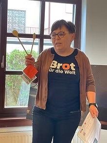 Eine Frau in einem blauen Shirt mit dem Slogan 'Brot für die Welt' steht drinnen und hält ein Paar Holzstäbe und ein Papier in den Händen, möglicherweise erklärt sie etwas.