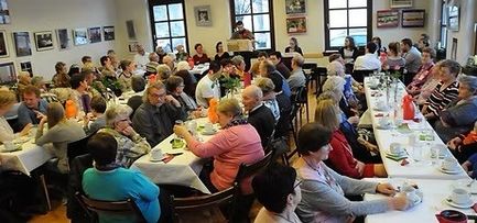 Eine Gruppe von Menschen sitzt an langen Tischen in einem Speisesaal, einige essen, andere unterhalten sich. Der Raum ist gut beleuchtet mit gerahmten Bildern an den Wänden.