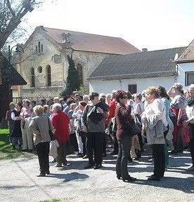Eine Gruppe von Menschen steht vor einem Gebäude mit einem roten Dach, einige tragen Brillen und Schuhe, und das Gebiet ist von Gras und Bäumen umgeben.