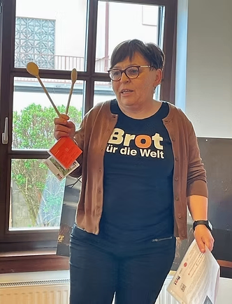 Eine Frau mit Brille und einem braunen Cardigan hält ein Paar Holzstäbe und eine kleine orangefarbene Schachtel mit den Worten 'Brot für die Welt' auf ihrem blauen Shirt. Sie steht vor einem Fenster mit Blick auf Bäume.