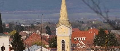 Luftaufnahme einer Stadt mit einem gelben Kirchturm, roten Dächern und einer entfernten Horizontlinie. Bäume und Gebäude sind sichtbar.