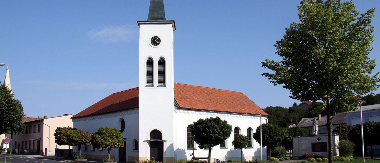 Eine weiße Kirche mit einem roten Dach und einem hohen Turm, der eine Uhr und einen Wetterhahn auf der Spitze hat. Die Kirche ist von Bäumen und Büschen umgeben.