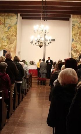 Eine Hochzeitszeremonie findet in einer Kirche statt. Der Priester liest aus einem Buch, und das Paar steht vor ihm. Die Gäste sitzen auf Bänken, und die Kirche ist mit Gemälden und einer Kronleuchter dekoriert.