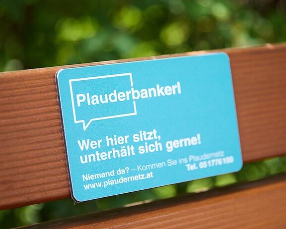 Ein blaues Schild mit weißem Text auf einer Holzbank, das 'Plauderbankerl' und 'Wer hier sitzt, unterhalt sich gerne!' mit einer Website und Telefonnummer anzeigt.