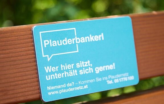 Ein blaues Schild mit weißem Text auf einer Holzbank, das 'Plauderbankerl' und 'Wer hier sitzt, unterhalt sich gerne!' mit einer Website und Telefonnummer anzeigt.