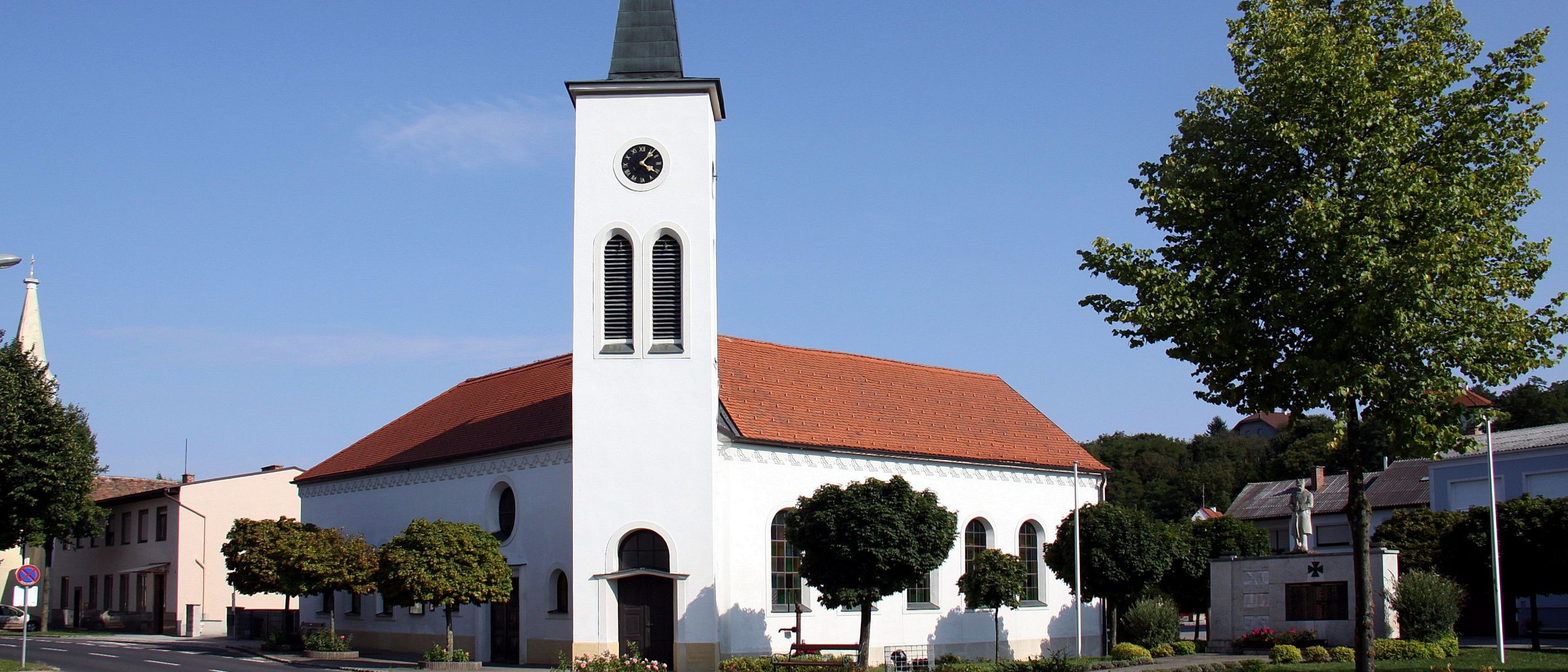 Eine weiße Kirche mit einem Kirchturm, einem roten Dach und einer Uhr. Sie hat Bogenfenster, eine braune Tür und ist von Grünpflanzen umgeben.