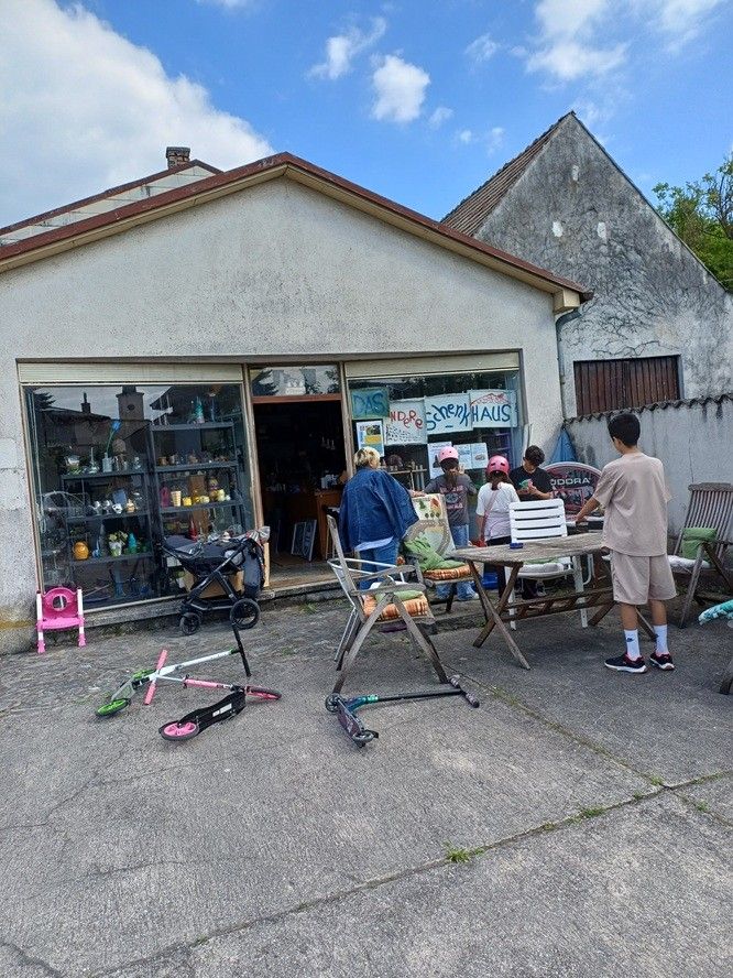 Eine Gruppe von Menschen steht vor einem Geschäft mit einem Schild, das 'Schneehaus' sagt. Ein Kinderwagen und einige Spielzeuge liegen auf dem Boden.