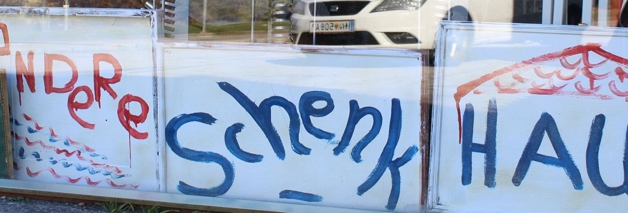 Ein weißes Schild mit blauer Graffiti, die 'Schenk' und ein rotes 'H' auf der rechten Seite liest, mit einem verschwommenen Spiegelbild eines weißen Autos und eines grünen Feldes im Hintergrund.