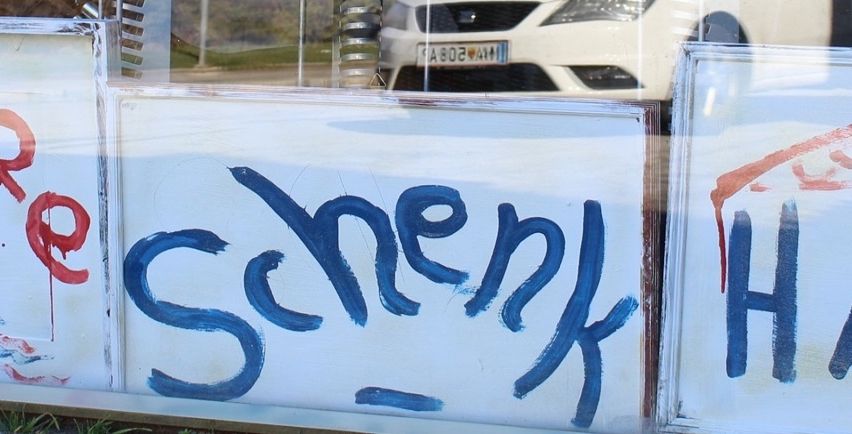 Ein weißes Schild mit blauer Graffiti, die 'Schenk' liest, ist in einem Fenster ausgestellt. Der Hintergrund zeigt eine verschwommene Ansicht eines weißen Autos und eines Grasbereichs.
