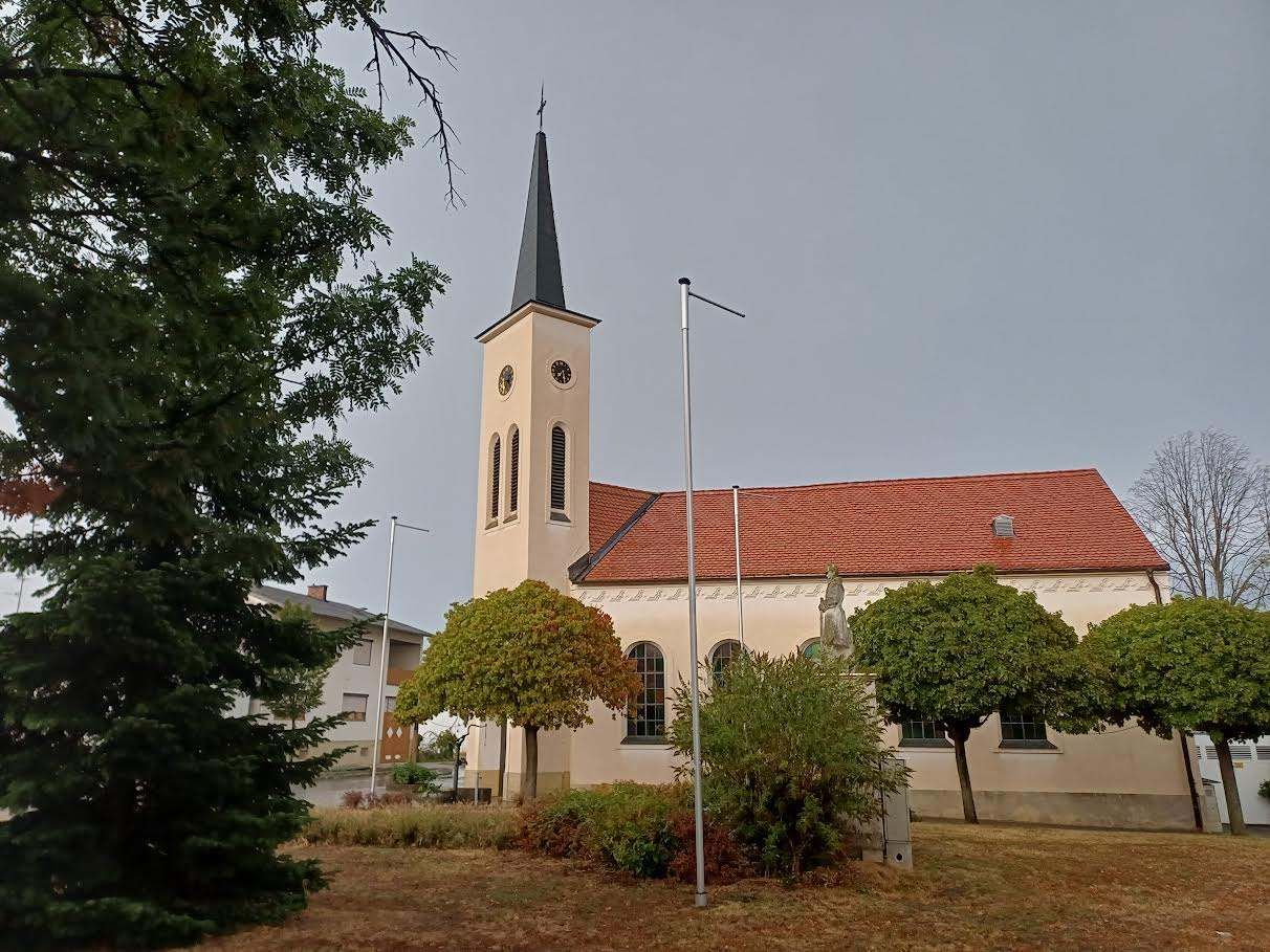 Eine traditionelle Kirche mit einem Turm und einem roten Dach, umgeben von Bäumen und Büschen. Das Gebäude hat eine Uhr und eine Statue.
