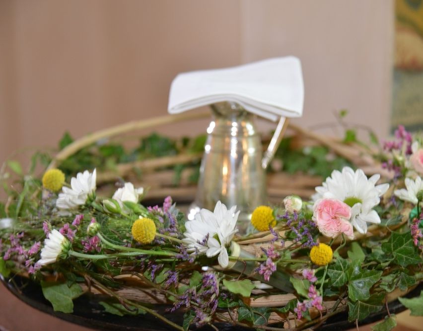 Ein Blumenkranz mit einer silbernen Kanne in der Mitte, geschmückt mit einer weißen Serviette oben. Die Blumen sind eine Mischung aus Weiß, Rosa und Gelb.