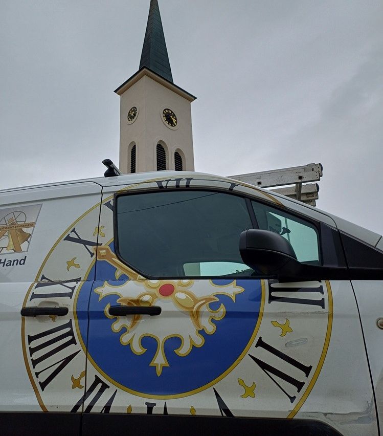 Ein weißer Van mit einem Logo, das römische Ziffern und einen Kirchturm im Hintergrund zeigt.