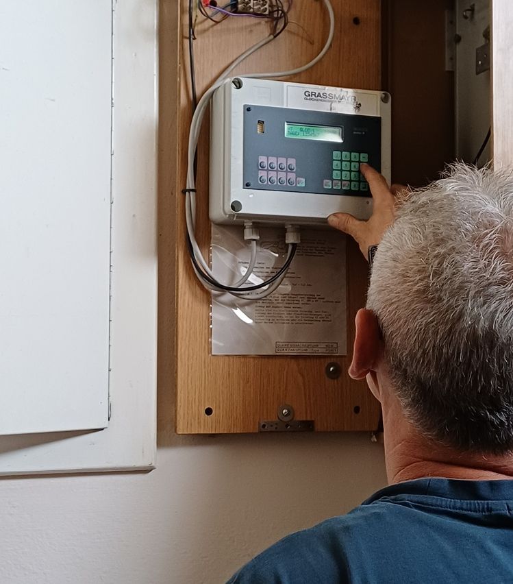 Ein Mann justiert ein Grassmayr-Gerät, das an einer Holzplatte montiert ist. Das Gerät hat ein digitales Display und Tasten.