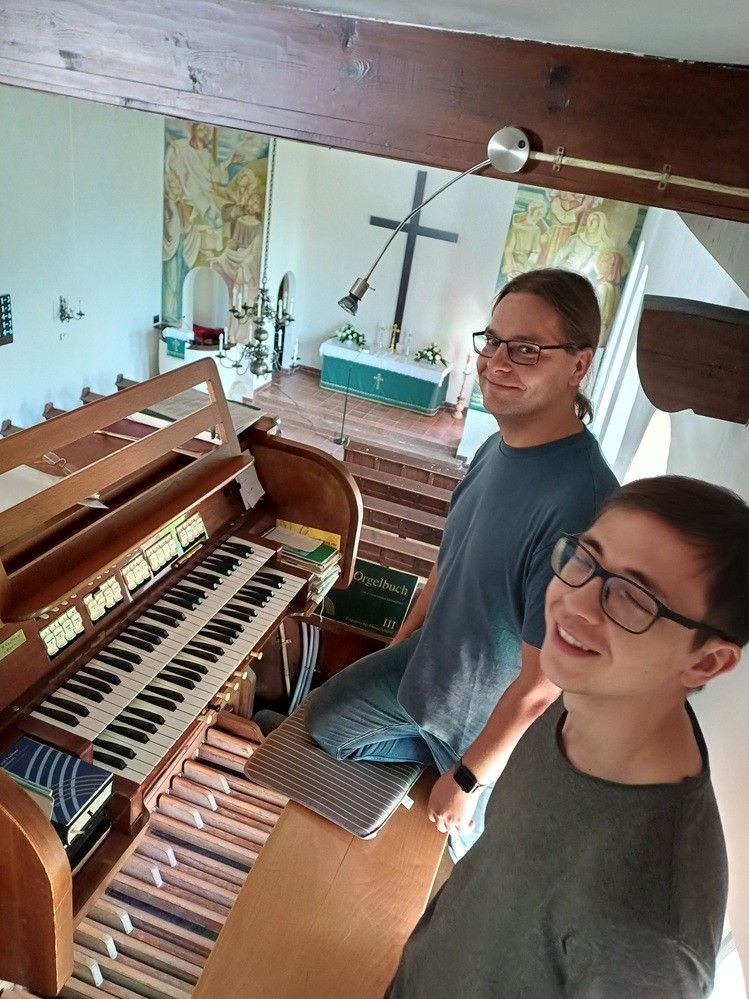 Zwei Männer lächeln und posieren für ein Foto in einer Kirche, mit einer großen Orgel im Vordergrund und einem Kreuz an der Wand hinter ihnen.