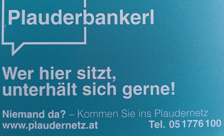 Blaues Schild mit weißem Text 'Plauderbaenkel' und einer Sprechblase mit 'Ver hier sitzt, unterhalt sich gerne!' darunter. Website und Telefonnummer sind ebenfalls angezeigt.