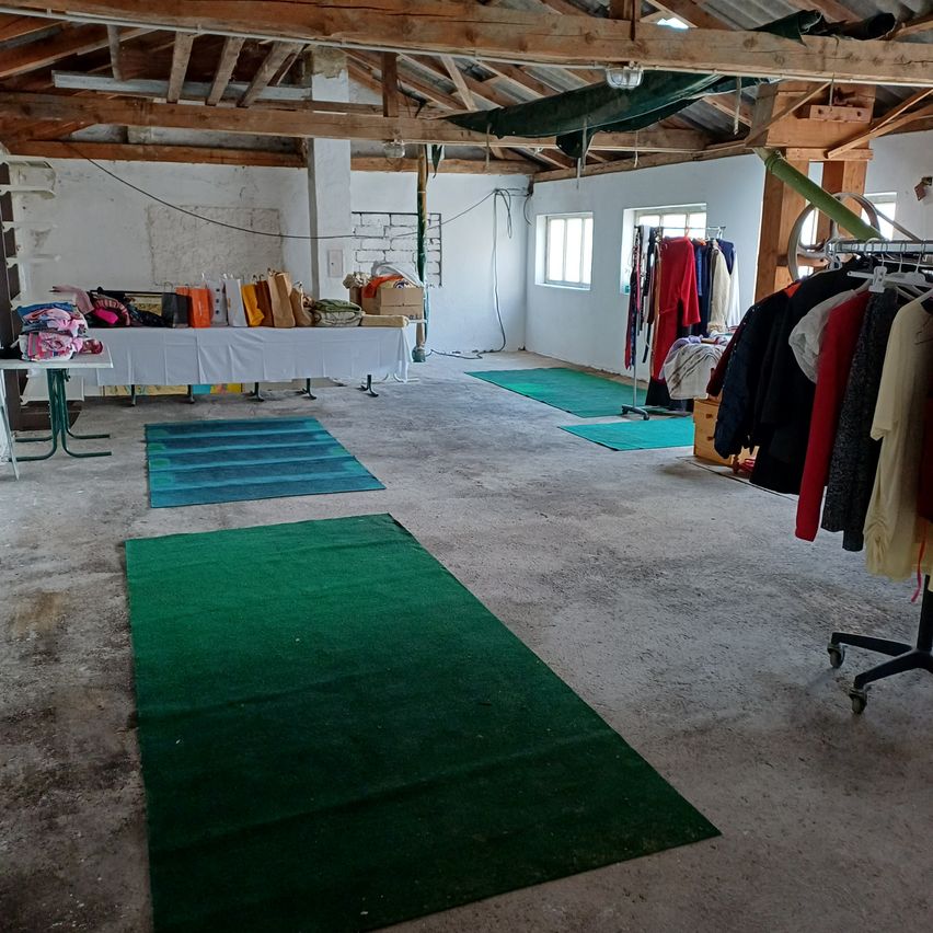 Ein leerer Raum mit Holzbalken und Betonboden. Es gibt drei grüne Teppiche auf dem Boden, einen Tisch mit verschiedenen Gegenständen und Kleidung an einem Kleiderständer. Der Raum hat zwei Fenster an der Wand.