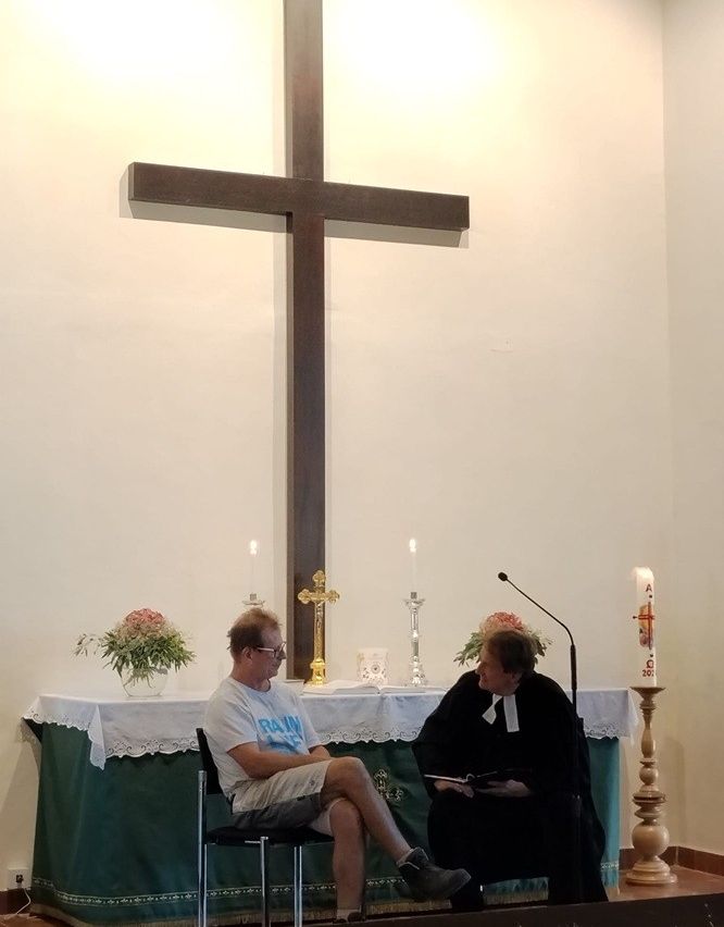 Ein Mann und eine Frau sitzen vor einem Kirchenaltar, mit einem Kreuz und Kerzen im Hintergrund.