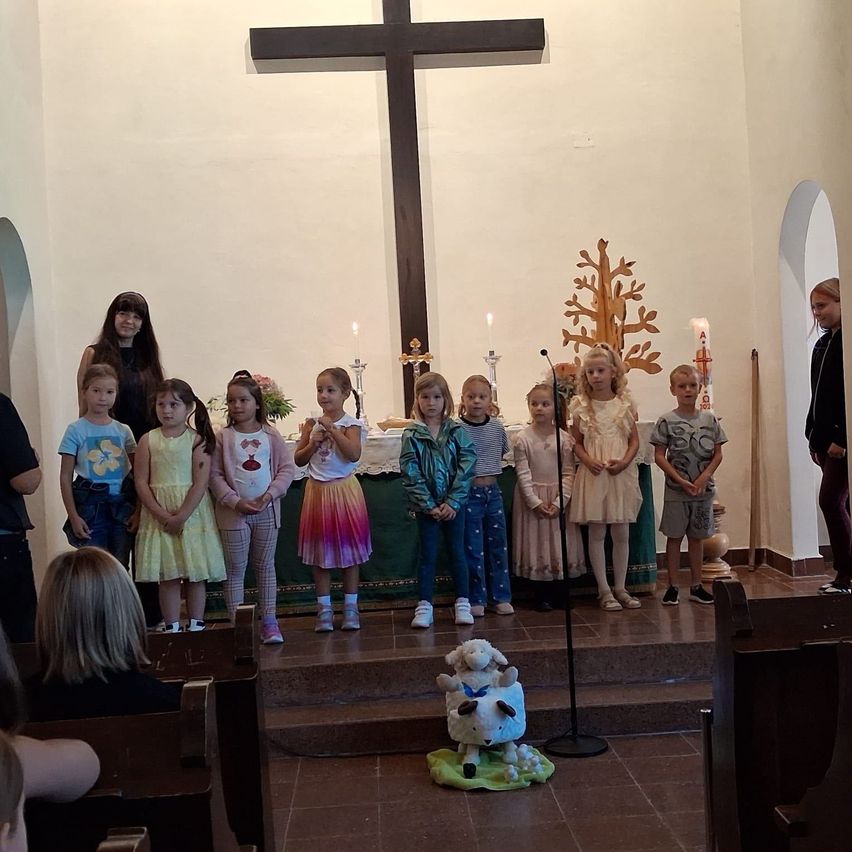 Eine Gruppe von Kindern steht vor einem Kirchenaltar mit einem Kreuz und Kerzen im Hintergrund. Sie sind in verschiedenen Outfits gekleidet und scheinen an einer religiösen Zeremonie teilzunehmen.