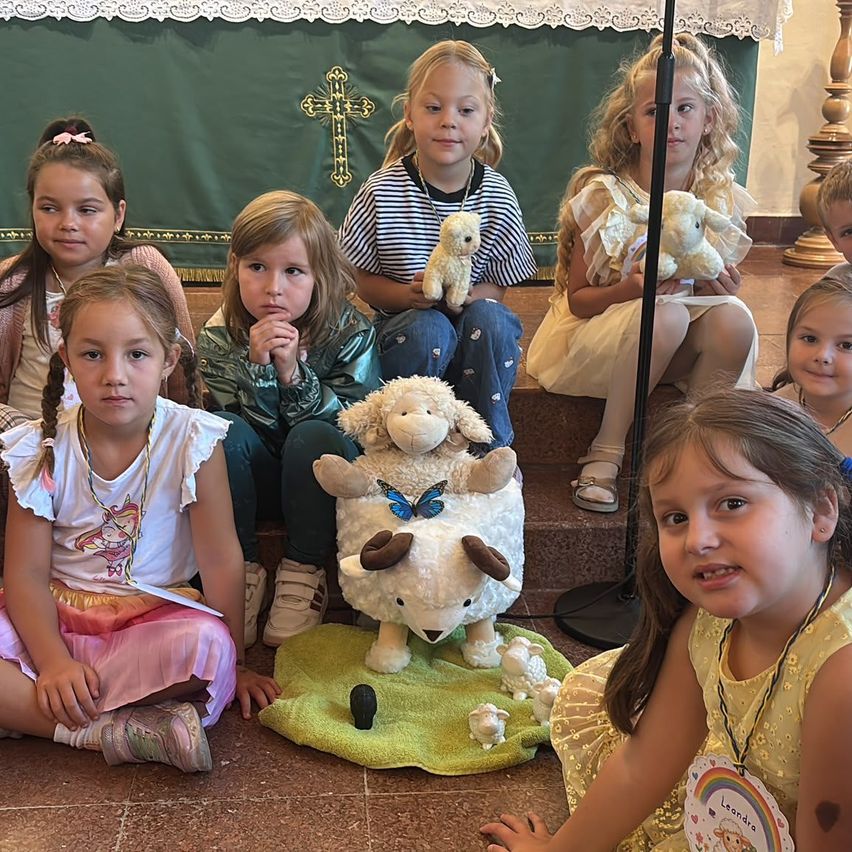 Eine Gruppe junger Mädchen sitzt auf dem Boden, einige halten Plüschtiere, und ein Mädchen hält ein großes Plüschschaf. Sie befinden sich in einem Raum mit grünem Hintergrund und Kreuzsymbol.