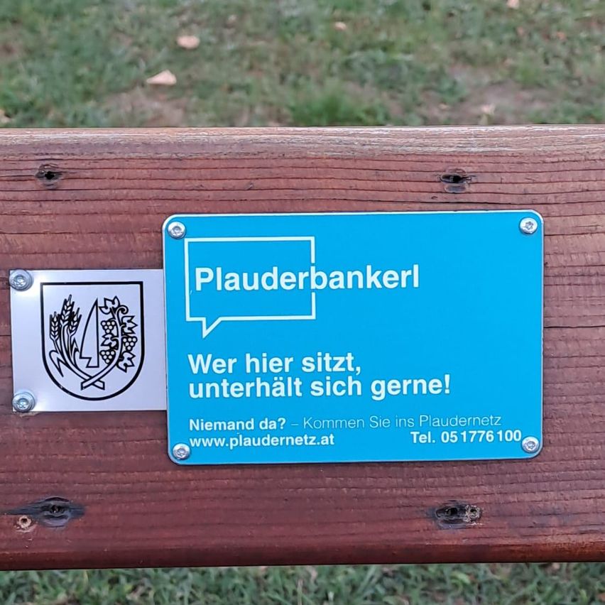 Ein blaues Informationsschild auf einer Holzbank mit dem Text 'Plaudernbankerl', einem Logo und einer Botschaft über das Sitzen und Genießen. Ein kleineres weißes Schild ist ebenfalls angebracht.