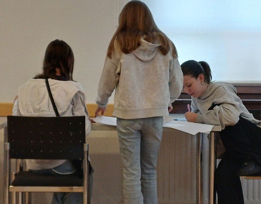 Drei Frauen in Freizeitkleidung sitzen an einem Schreibtisch, eine schreibt auf Papier. Die Frau in der Mitte trägt eine Kapuzenjacke.