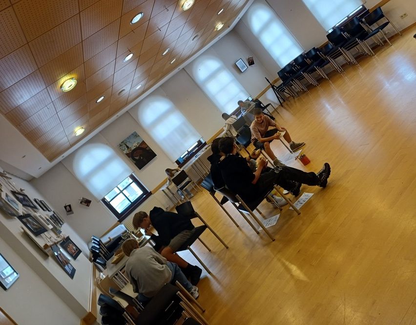 Eine Gruppe von Menschen sitzt auf Stühlen in einem Raum mit Holzböden und großen Fenstern. Eine Person benutzt einen Laptop, während andere in Gespräche vertieft sind. An der Wand hängen gerahmte Bilder und im Hintergrund sind Stühle ordentlich aufgestellt.