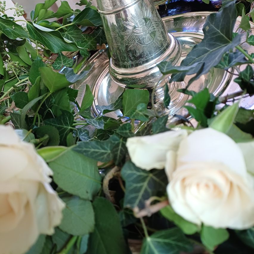 Ein Nahaufnahme von weißen Rosen und grünen Blättern mit einer silbernen Glocke in der Mitte. Die Glocke ist kunstvoll mit einer Engelsfigur verziert.