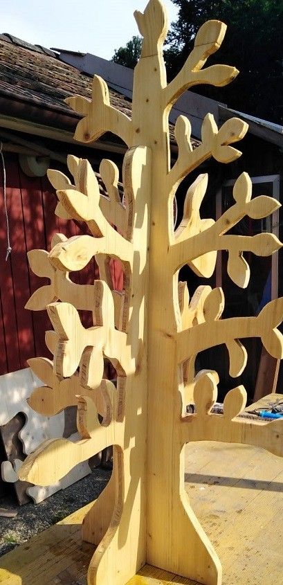 Eine hölzerne Baumskulptur steht auf einer Holzfläche mit kunstvollen Blattdesigns in ihren Ästen. Der Baum befindet sich vor einem roten Gebäude.