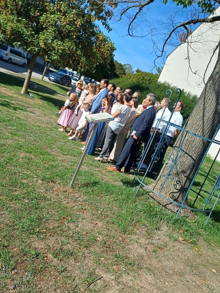 Eine Gruppe von Menschen, einschließlich Kinder, steht in einem Park, möglicherweise für eine Zeremonie, mit einem Baum und einem weißen Gebäude im Hintergrund.