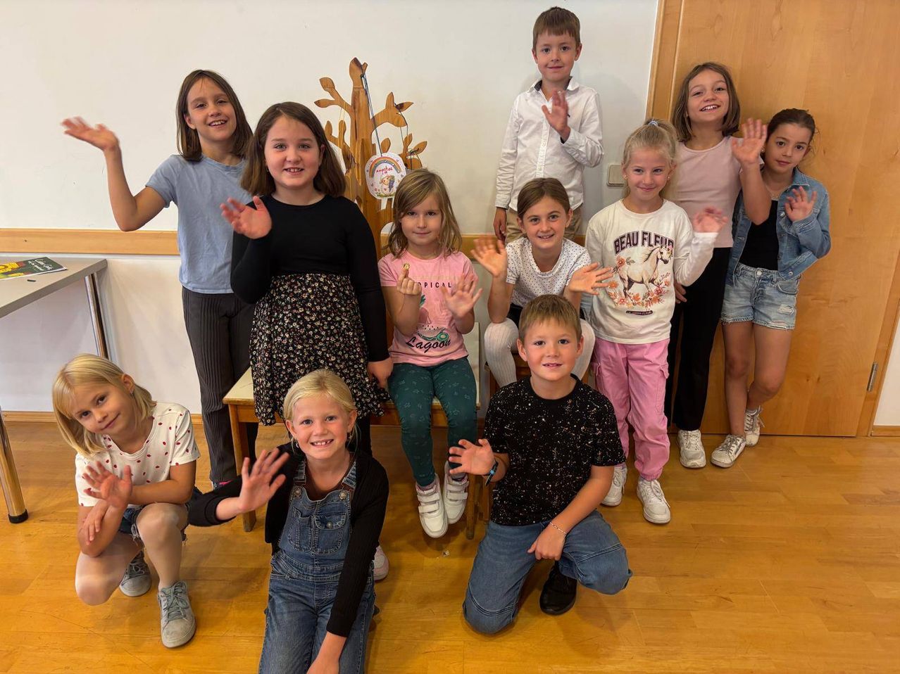 Eine Gruppe von Kindern, sowohl Jungen als auch Mädchen, posiert für ein Foto im Klassenzimmer. Sie alle lächeln und machen Handgesten. Der Raum hat Holzböden und weiße Wände.