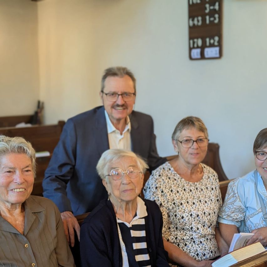 Vier ältere Personen, zwei Männer und zwei Frauen, sitzen auf einer Kirchenbank und lächeln für ein Foto. Der Mann in der Mitte trägt einen Anzug und eine Brille.