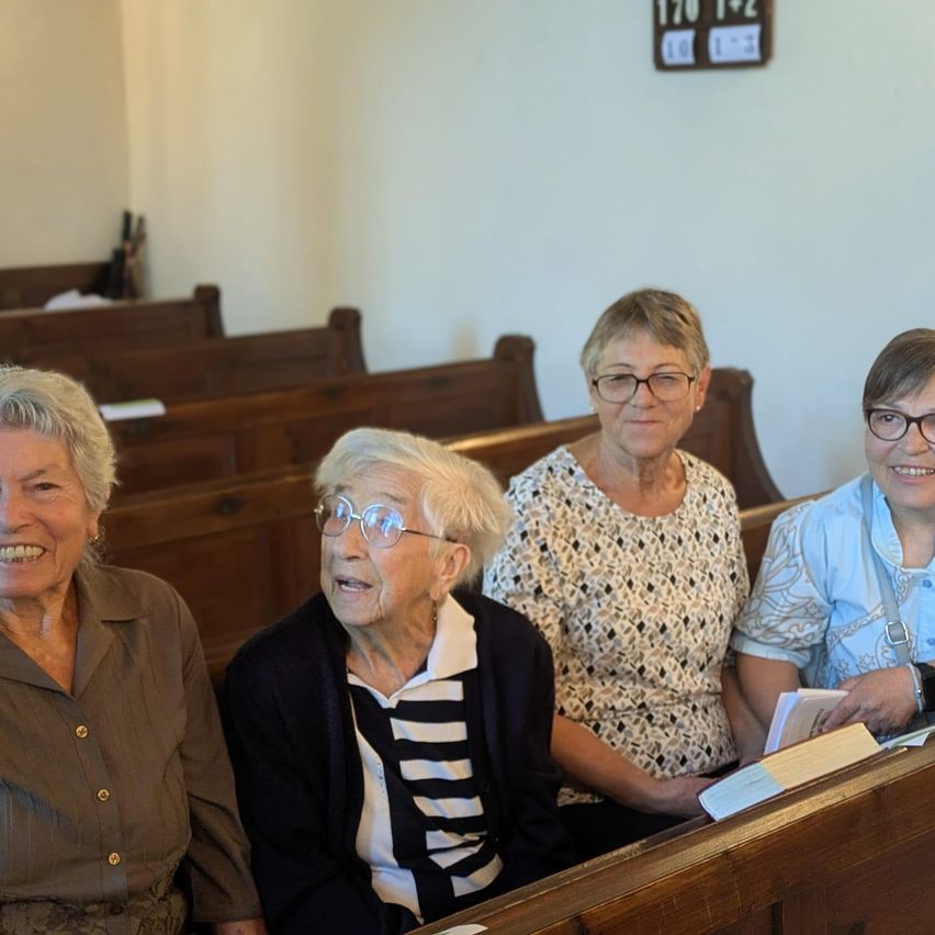 Vier ältere Frauen sitzen in einer Kirchenbank, lächeln und tragen Brillen. Die Frau auf der linken Seite trägt ein braunes Blusenhemd, die mittlere ein schwarz-weiß gestreiftes Hemd und die beiden auf der rechten Seite gemusterte Blusen. Hinter ihnen ist eine Wand mit einer digitalen Uhr.