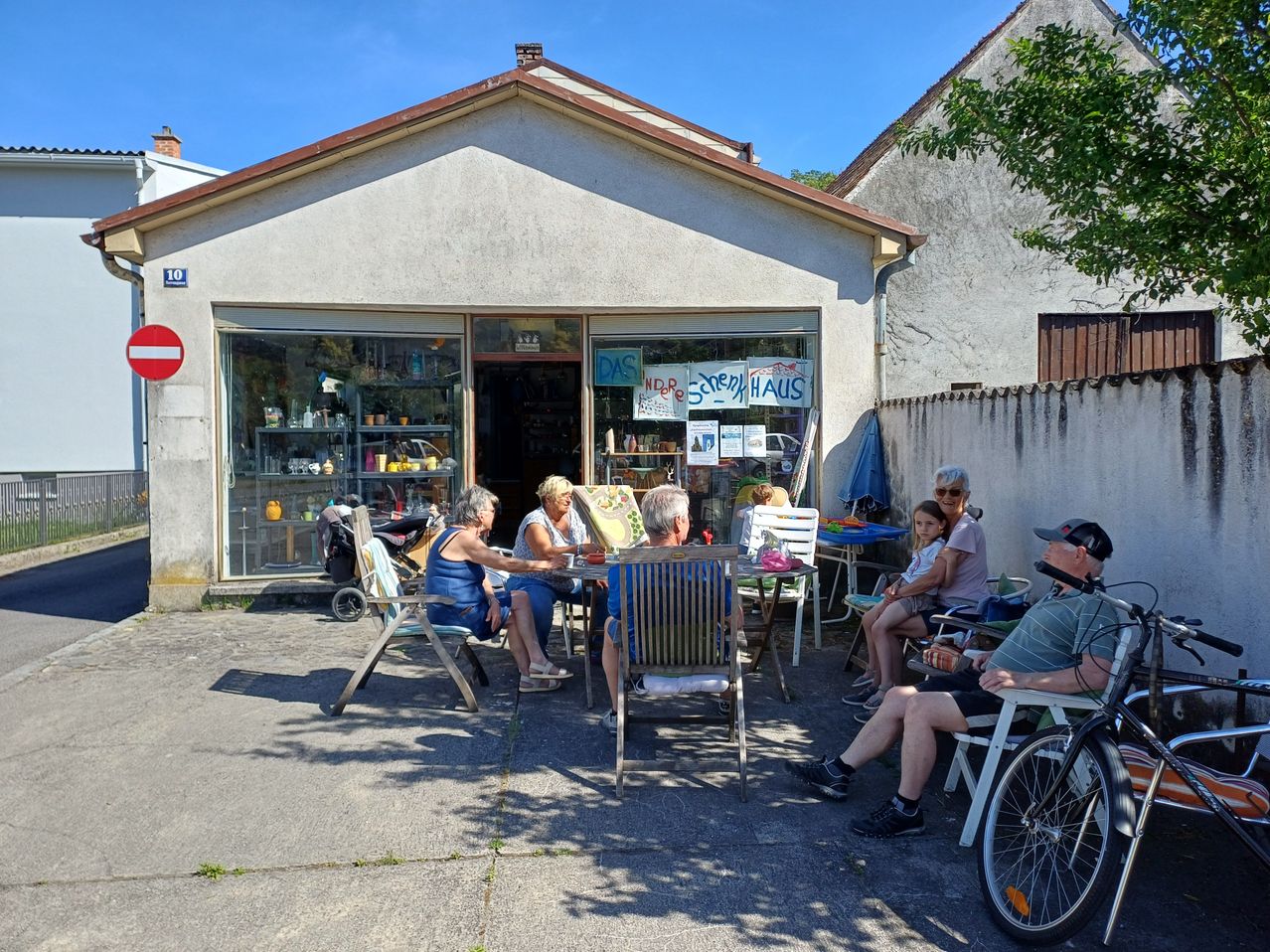Eine Gruppe von Menschen versammelt sich vor einem Geschäft. Sie sitzen um einen Tisch herum. Das Geschäft hat ein Schild mit der Aufschrift 'DAS NBre SCHenk HAUS'. Das Gebäude hat eine Glastür mit Regalen im Inneren. Vor ihnen steht ein Fahrrad geparkt.