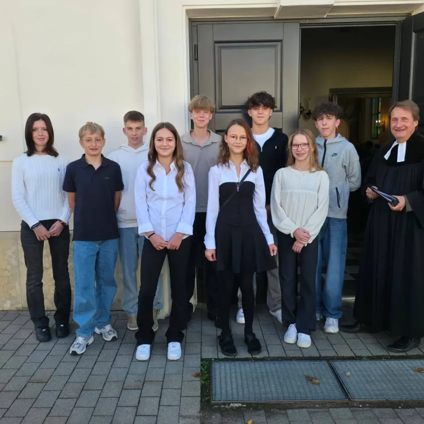 Eine Gruppe junger Menschen und ein erwachsener Mann, in verschiedenen Outfits gekleidet, stehen vor einem Gebäude. Der Erwachsene lächelt und hält ein Buch.