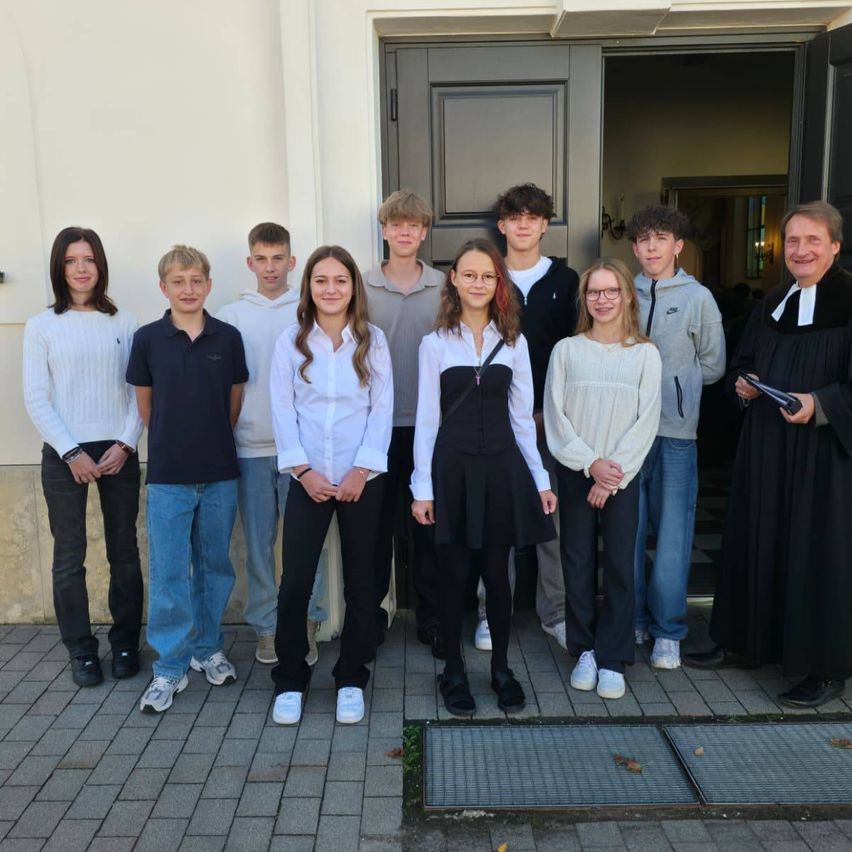 Eine Gruppe junger Menschen und ein erwachsener Mann, in verschiedenen Outfits gekleidet, stehen vor einem Gebäude. Der Erwachsene lächelt und hält ein Buch.