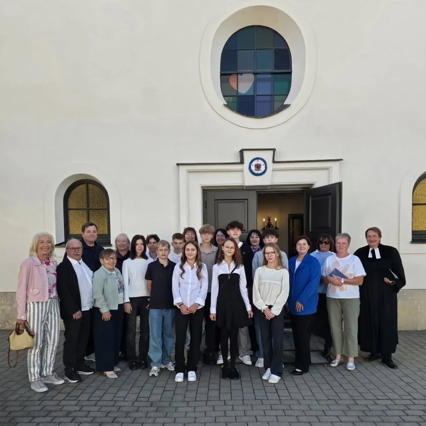 Eine Gruppe von Menschen, darunter Erwachsene und Kinder, steht vor einem weißen Gebäude mit Buntglasfenstern. Sie alle lächeln und posieren für ein Foto.