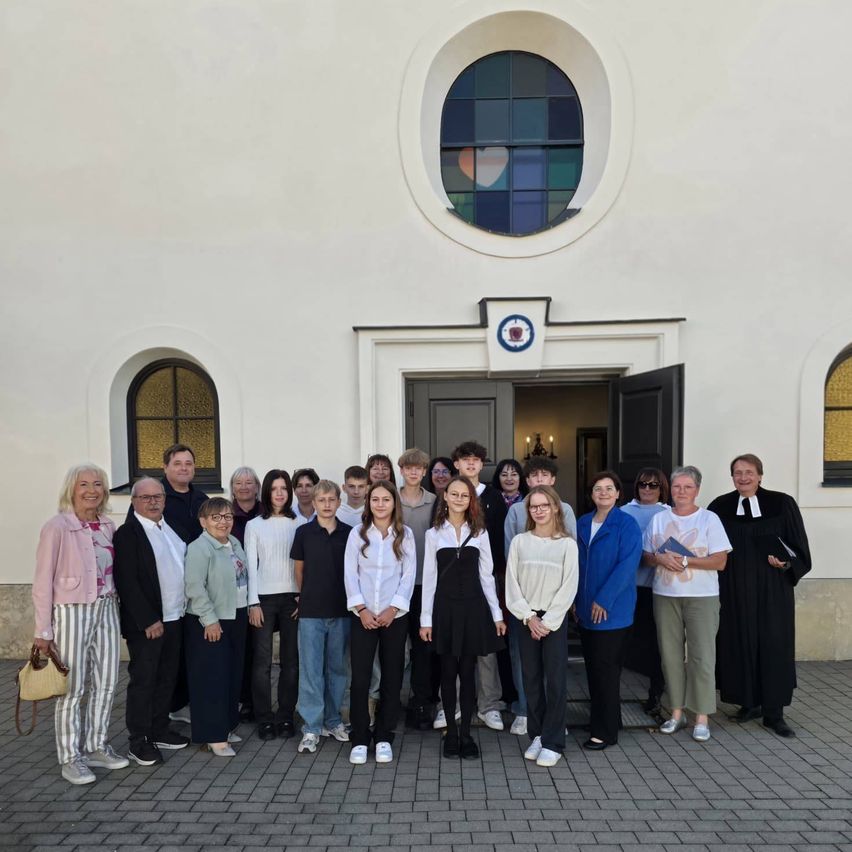 Eine Gruppe von Menschen, darunter Erwachsene und Kinder, steht vor einem weißen Gebäude mit Buntglasfenstern. Sie alle lächeln und posieren für ein Foto.