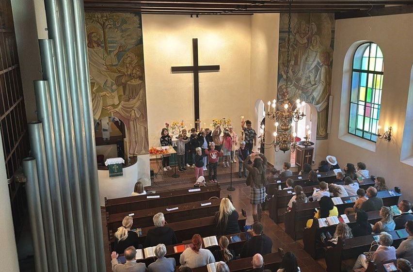 Ein Kircheninneres mit einem Kreuz am Altar, Buntglasfenstern und einer Gemeinde, die auf Kirchenbänken sitzt.