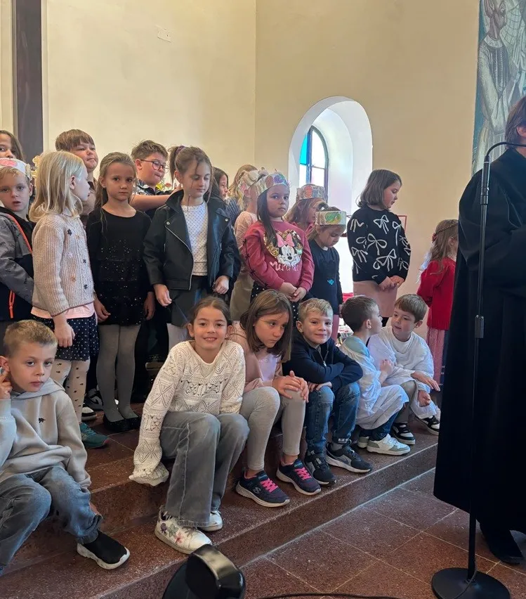 Eine Gruppe von Kindern in einer Kirche, einige sitzen auf Stufen, andere stehen, alle tragen Winterkleidung und einige tragen Partyhüte. Eine Person in einem schwarzen Gewand steht rechts, möglicherweise ein Priester.