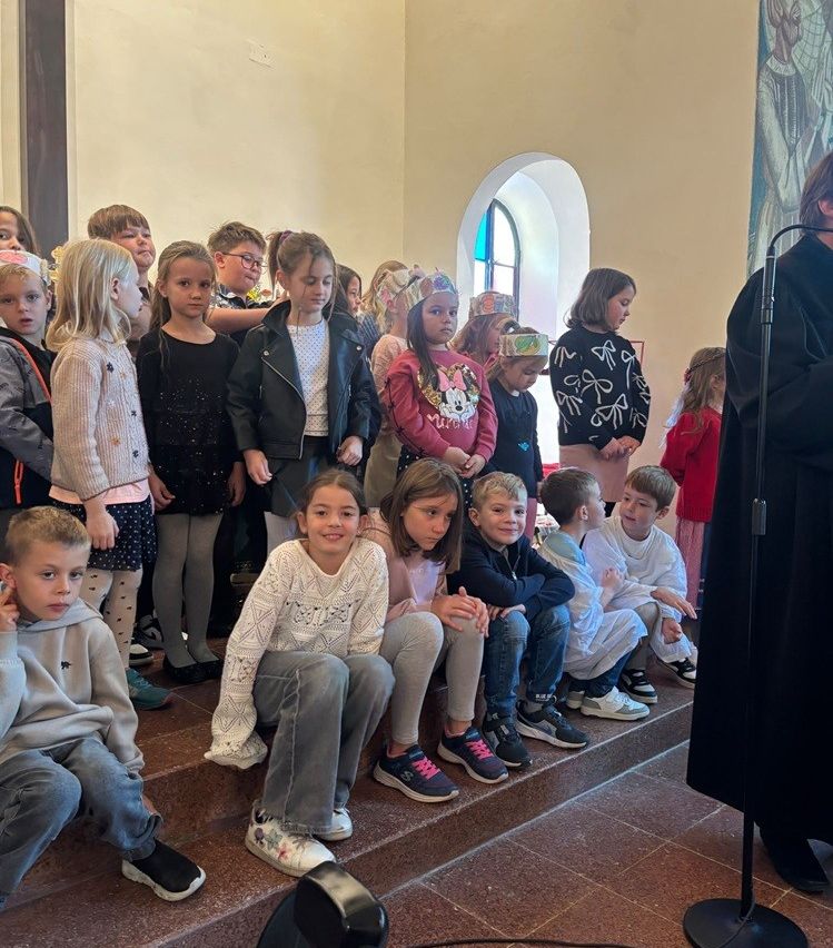 Eine Gruppe von Kindern in einer Kirche, einige sitzen auf Stufen, andere stehen, alle tragen Winterkleidung und einige tragen Partyhüte. Eine Person in einem schwarzen Gewand steht rechts, möglicherweise ein Priester.