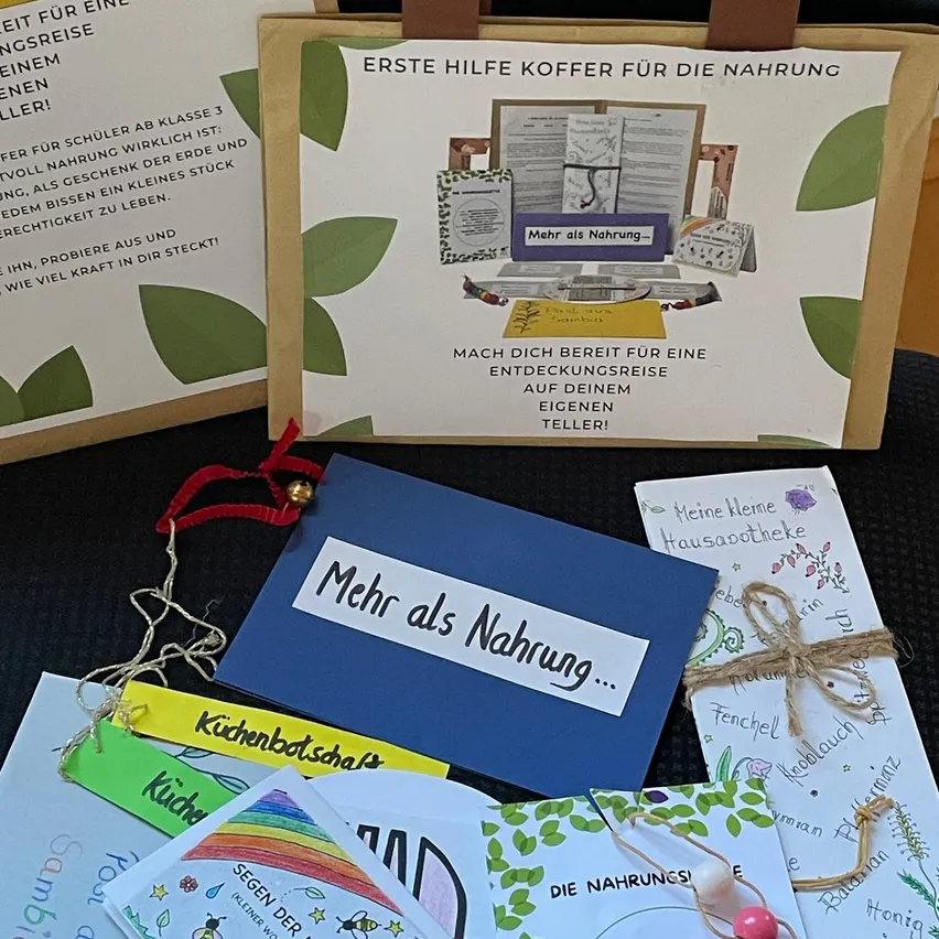 Eine Sammlung von Lernmaterialien für Kinder, einschließlich einer blauen Mappe mit der Aufschrift 'Mehr als Nahrung', einem braunen Papierbeutel mit grünem Blattdesign und verschiedenen bunten Karten und Broschüren.