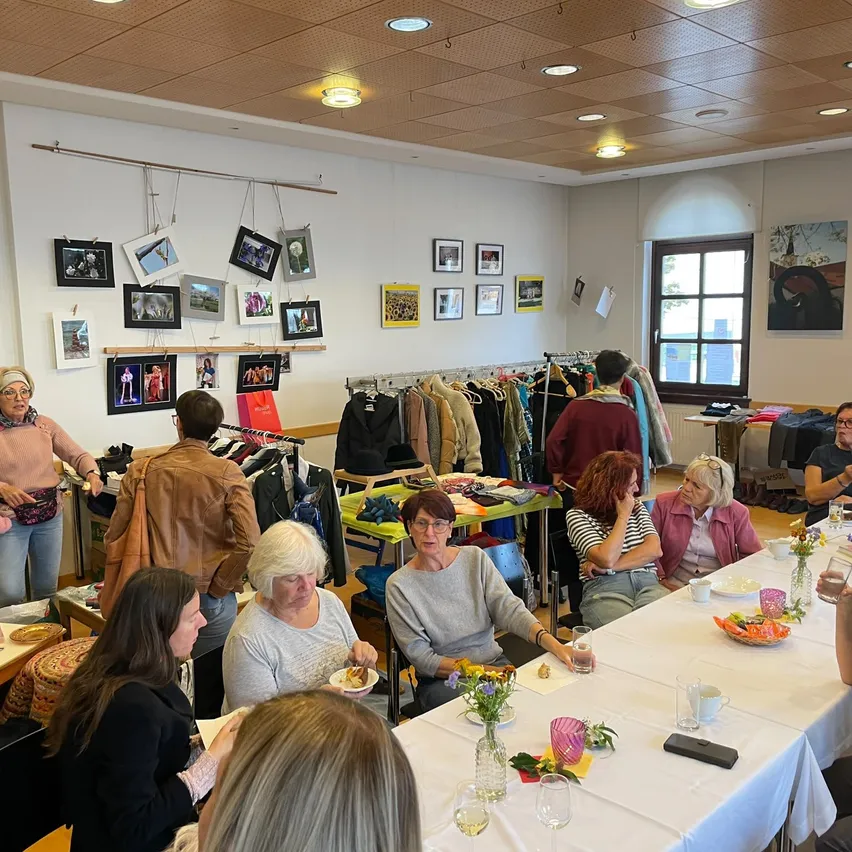 Eine Gruppe von Frauen sitzt an einem langen Tisch in einem Raum. Sie sitzen und stehen, einige halten Gläser, und es hängen Bilderrahmen an der Wand.