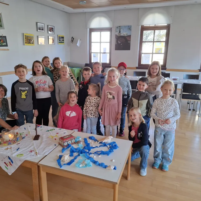 Eine Gruppe von Kindern und einer erwachsenen Frau versammelt sich in einem Raum, möglicherweise für eine Feier. Die Kinder stehen vor einem Tisch mit einem Kuchen und anderen Dekorationen. Der Raum hat weiße Wände und Holzböden.